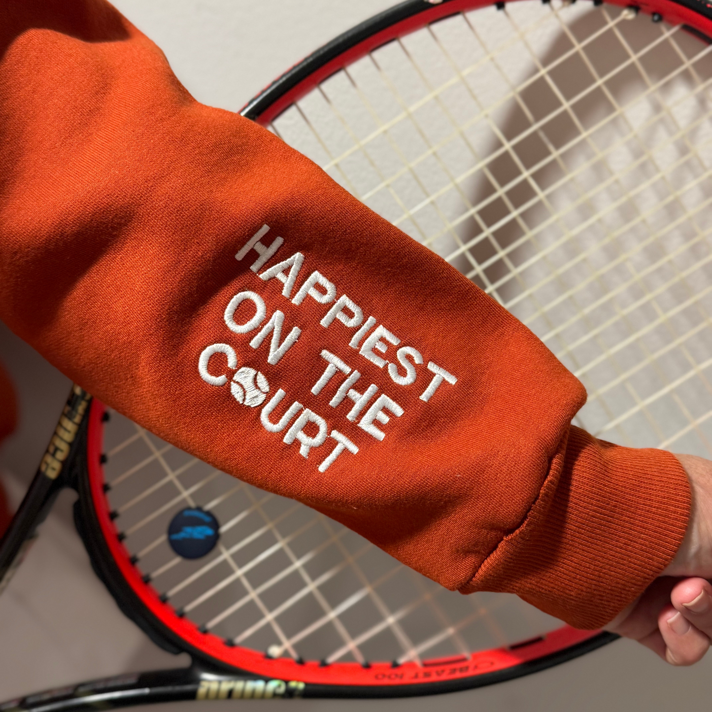 Orange sweatshirt with 'Happiest on the Court' text held by a person with a tennis racket.
