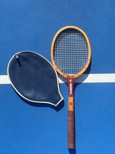 A vintage BORG tennis racquet with a yellow-orange frame and brown grip, shown alongside its black teardrop-shaped rubber case on a blue tennis court background.
