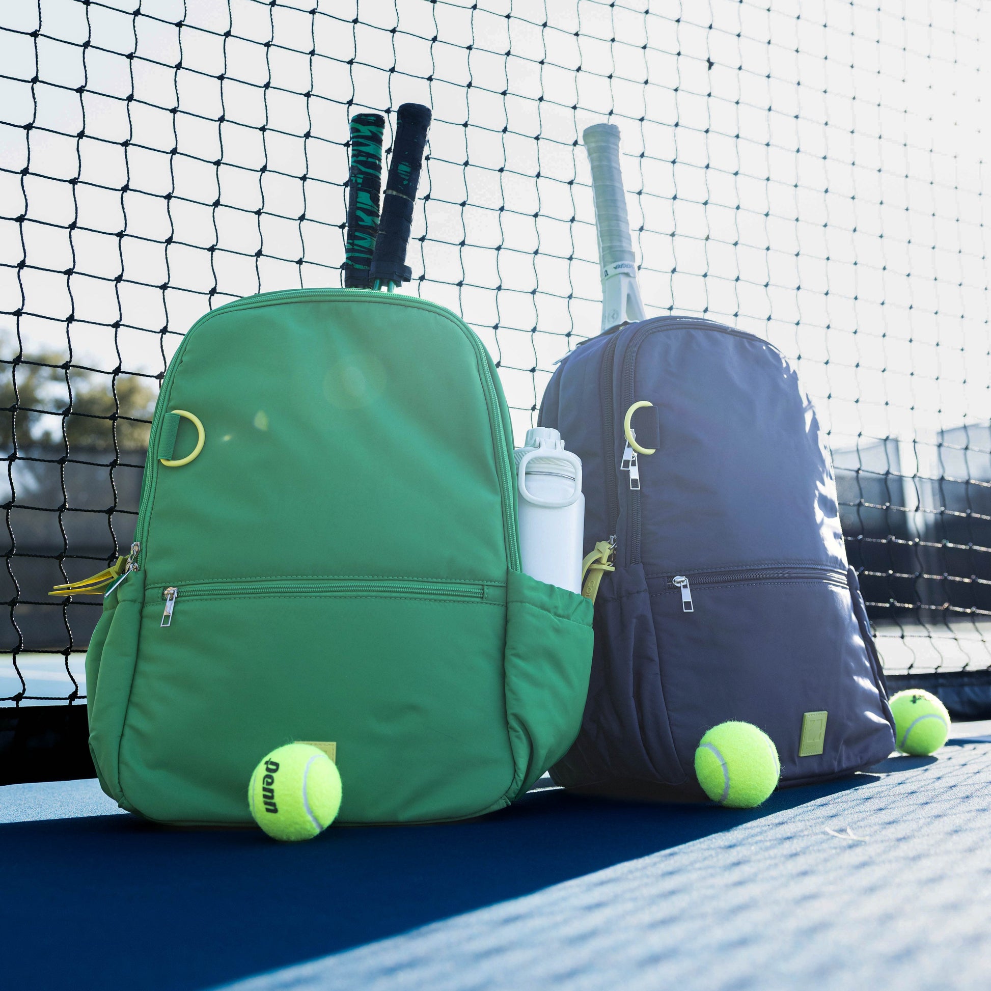 Two backpacks on a tennis court with tennis balls and rackets.