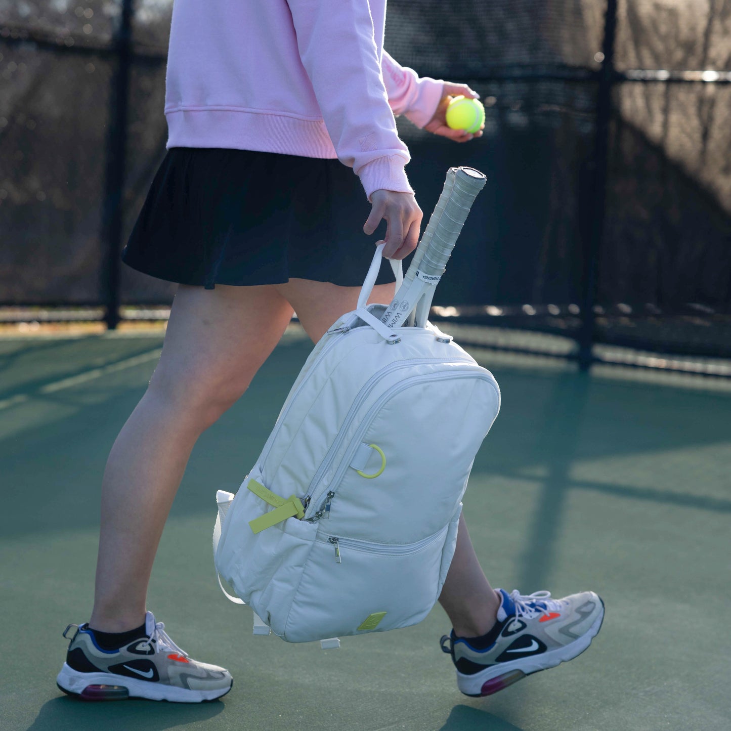 Person holding a tennis racket and ball, walking on a tennis court with a white backpack.