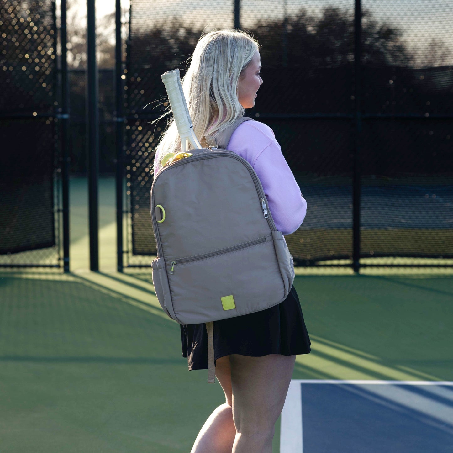 Woman on a tennis court wearing a gray backpack with a tennis racket.