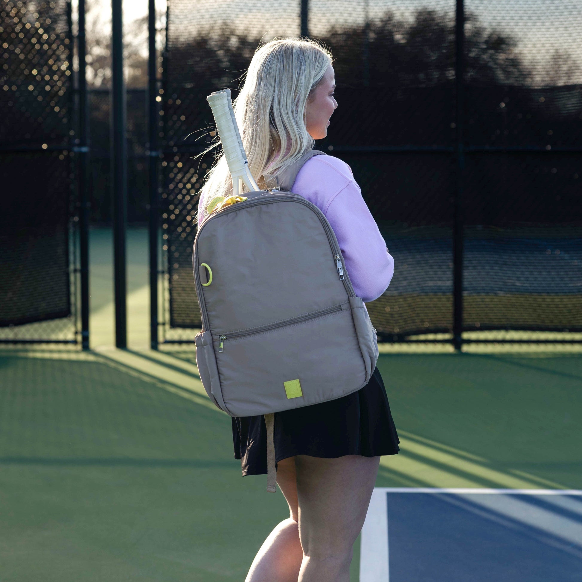 Woman on a tennis court wearing a gray backpack with a tennis racket.