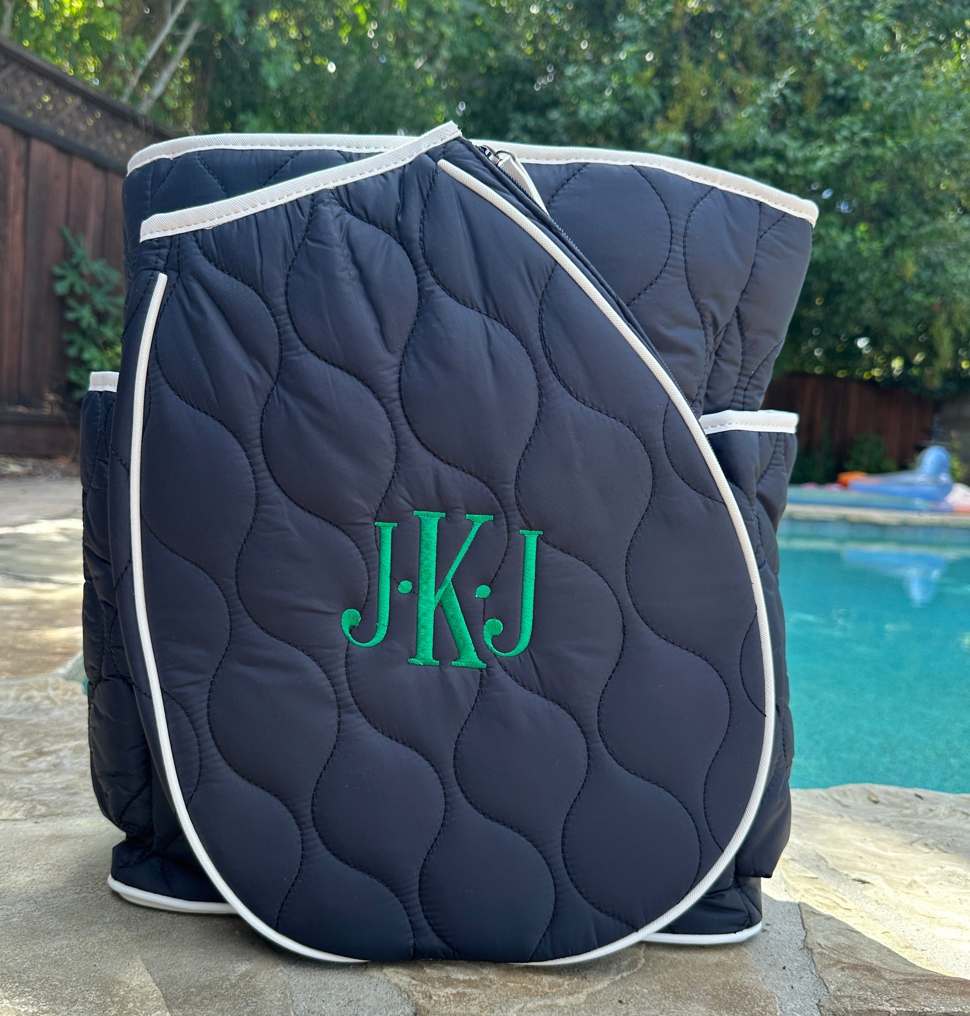 Navy quilted tennis bag with green monogram 'J.K.J' by a poolside.
