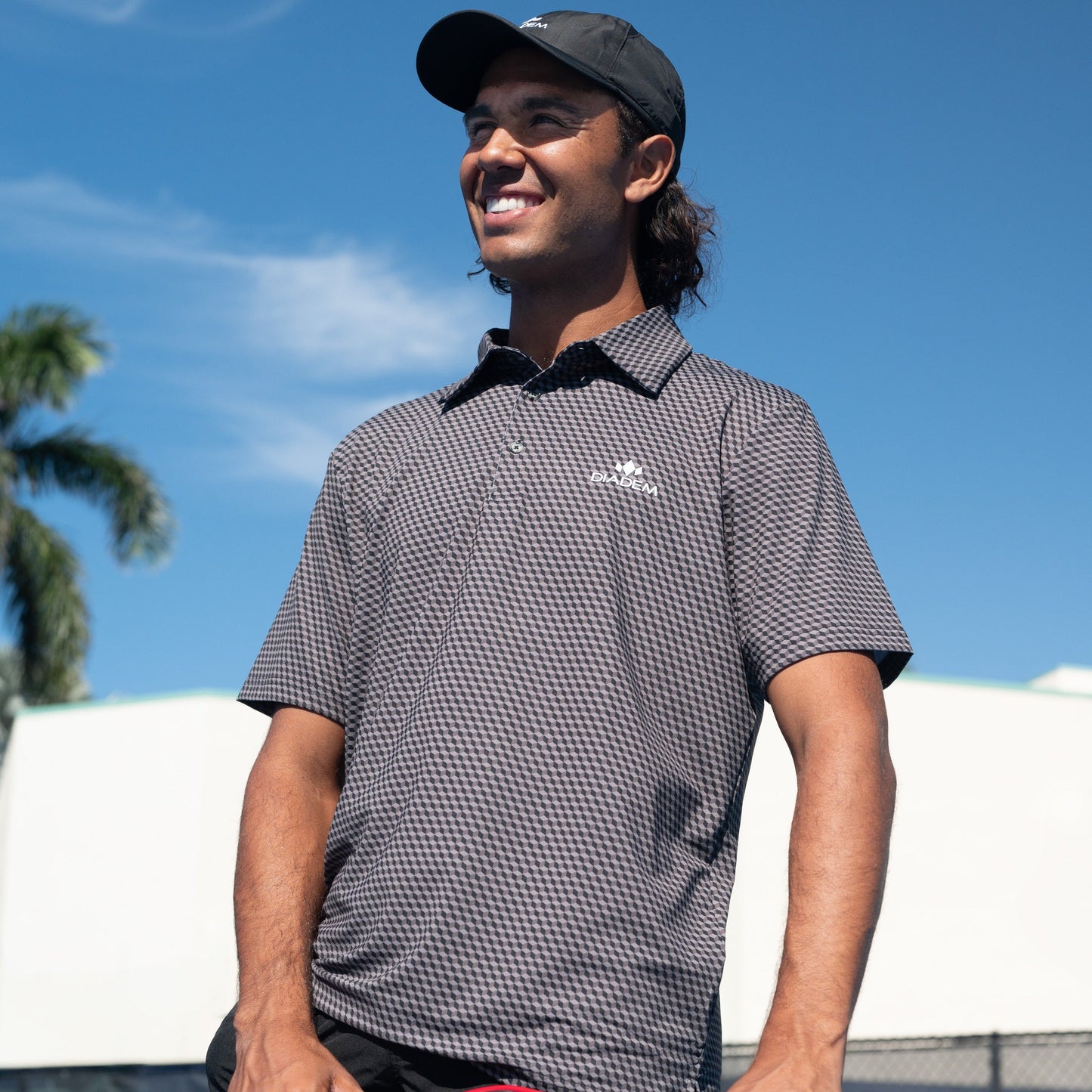 Man wearing a checkered polo shirt with a logo outdoors against a blue sky.
