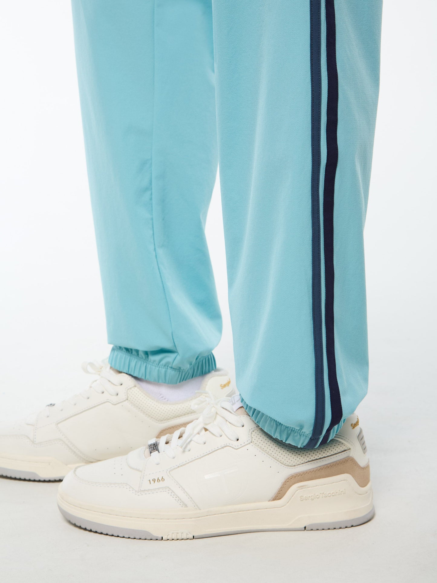 Light blue tracksuit pants with a dark stripe worn with white sneakers on a light background