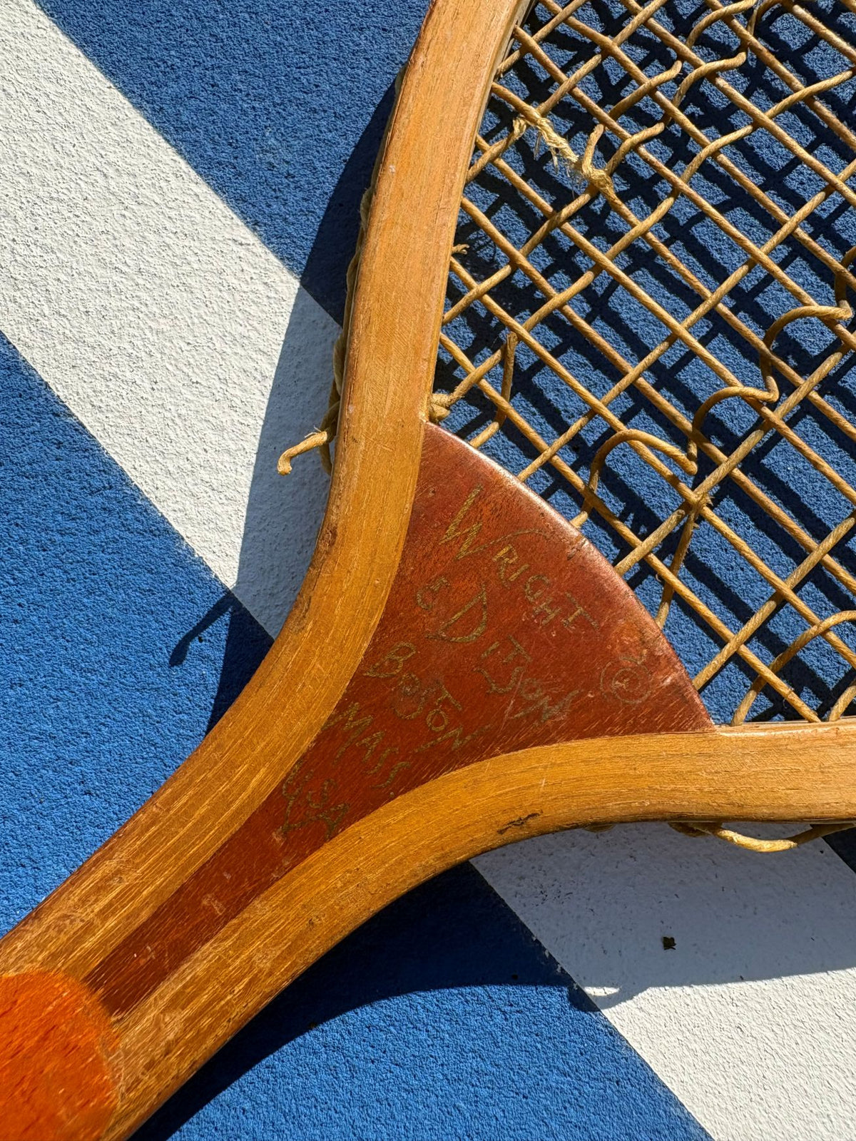 Close-up of a vintage wooden tennis racket with gold-colored strings, resting on a blue and white striped tennis court surface.