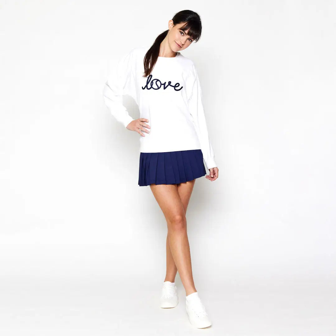 Woman wearing a white tennis  sweatshirt with 'love' text and a blue pleated skirt on a white background