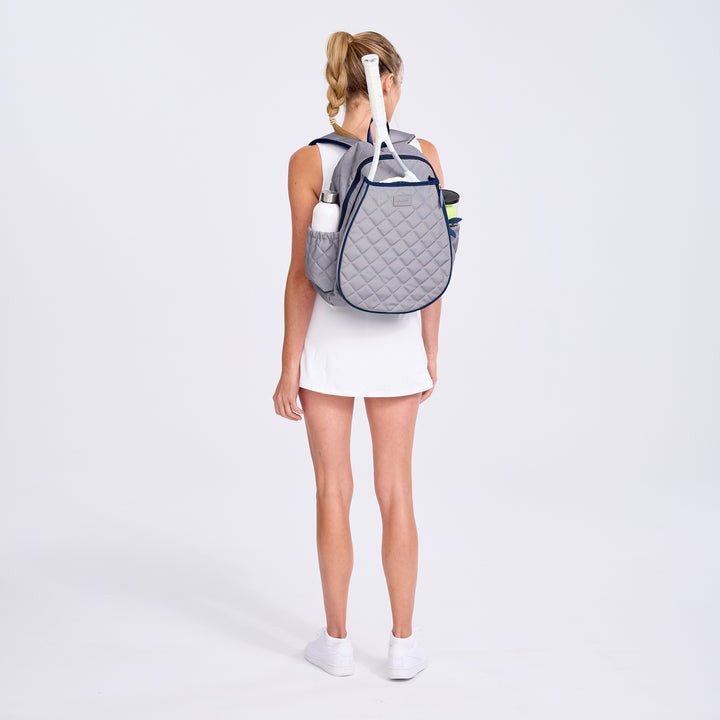 Woman wearing a gray quilted tennis backpack with sports equipment on a white background