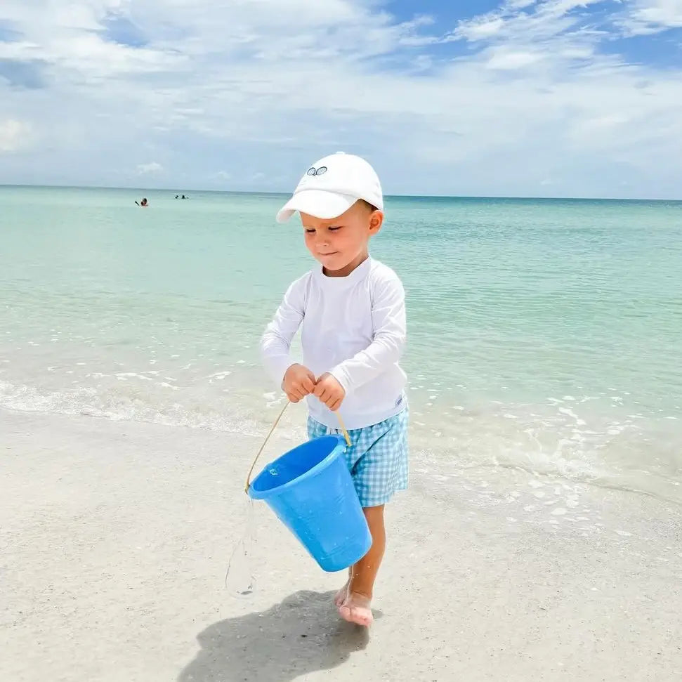 Child playing on a beach with a blue bucket, wearing a white cap and checkered shorts.