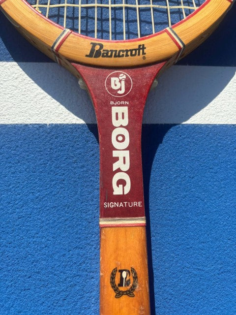 A close-up of a vintage Borg tennis racket with a wooden handle and red throat, resting on a blue and white tennis court surface.