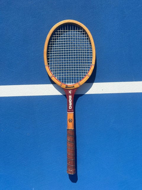 A vintage-style Borg tennis racket with a yellow-orange frame and brown grip, laid diagonally on a blue tennis court with a white boundary line.