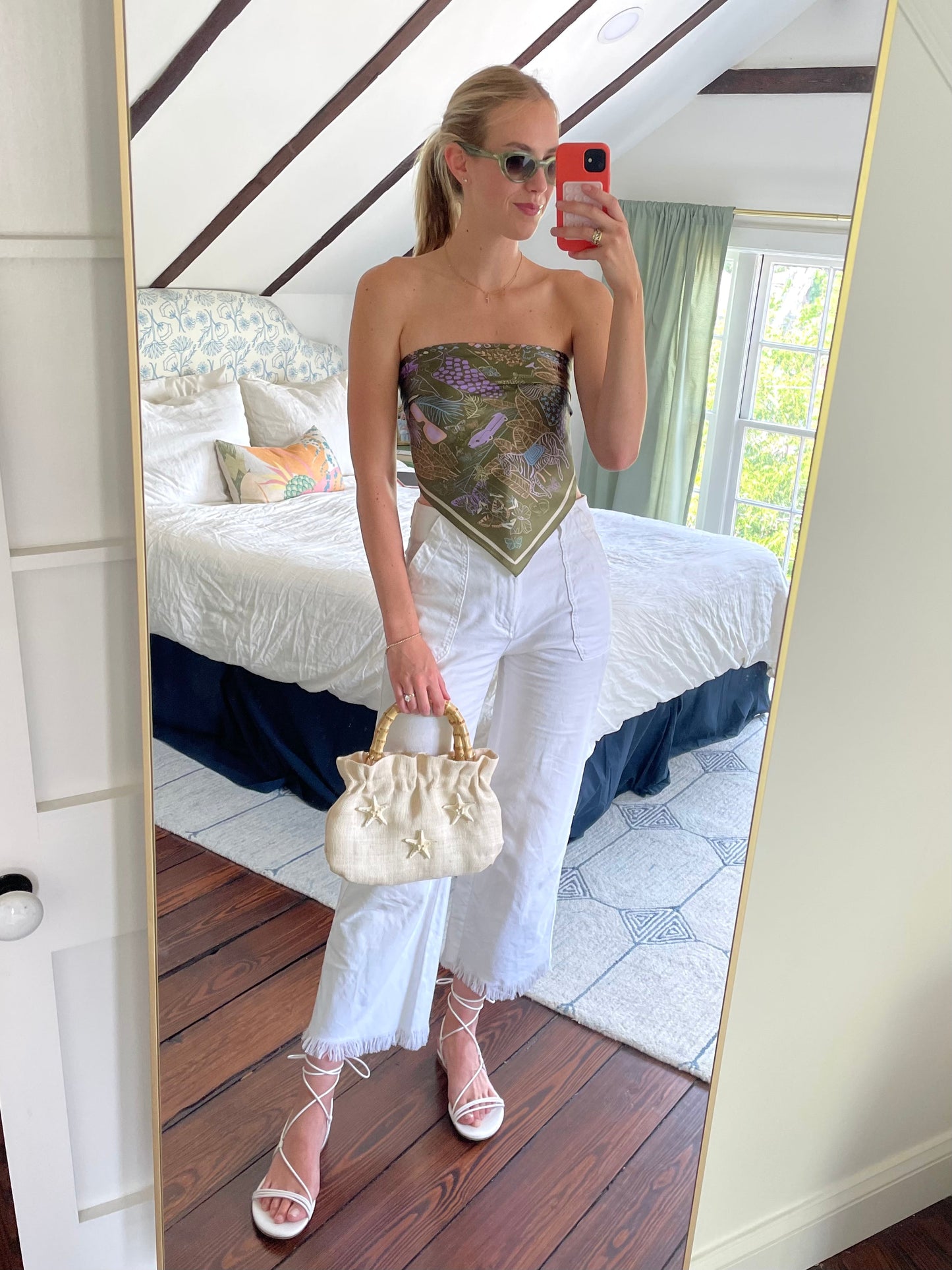 Woman taking a mirror selfie wearing a strapless top and white pants, holding a beige handbag.