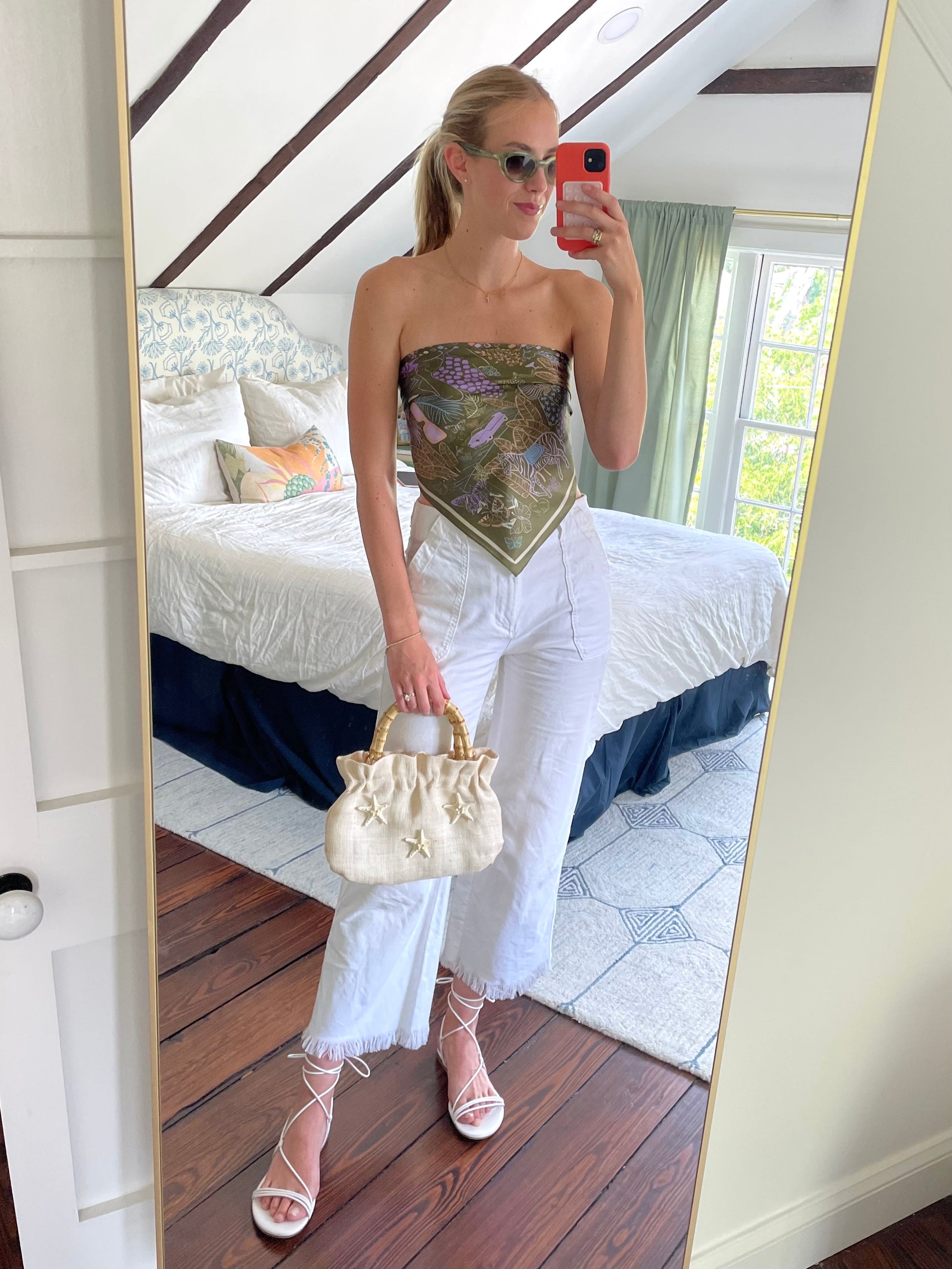 Woman taking a mirror selfie wearing a strapless top and white pants, holding a beige handbag.