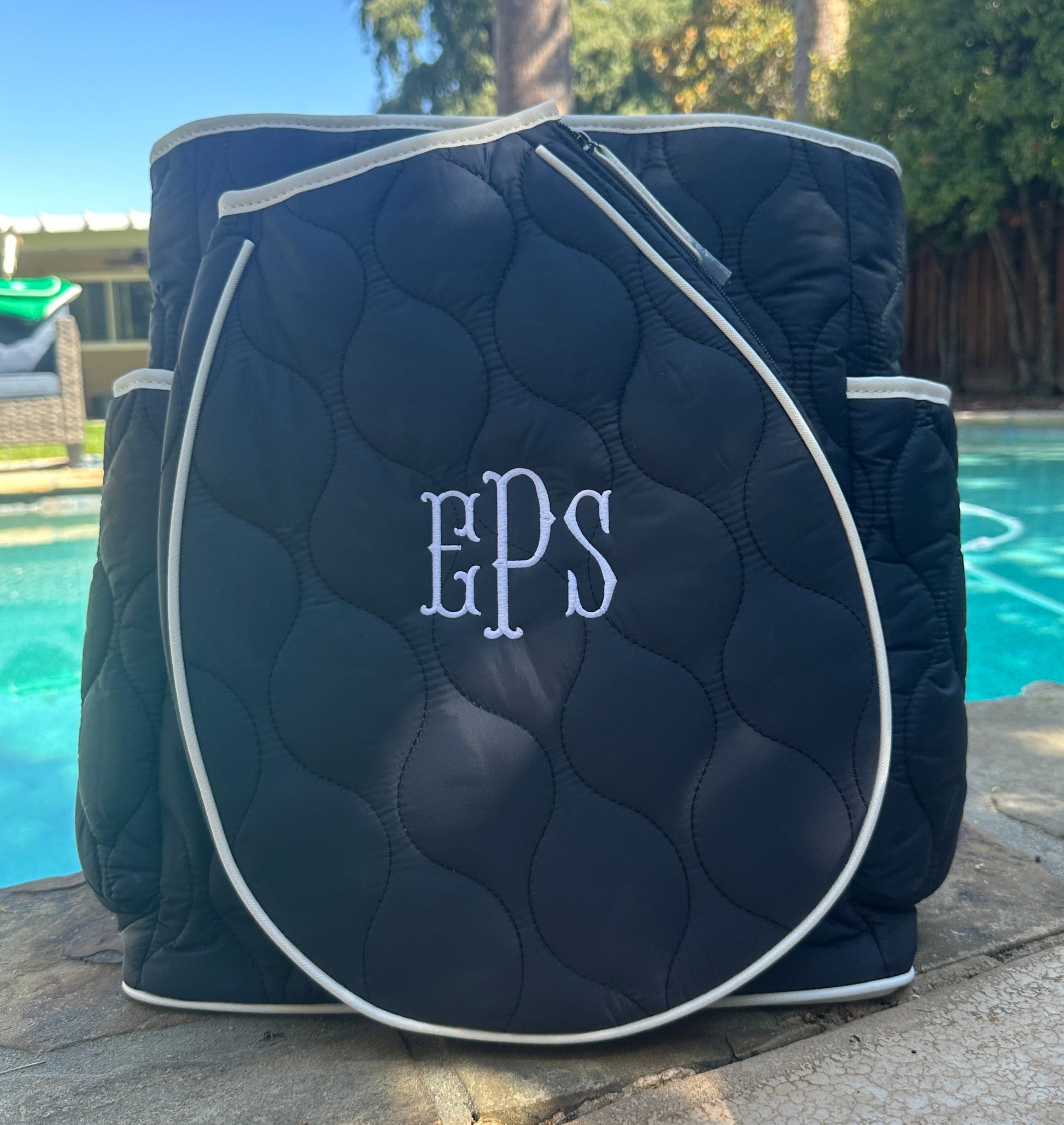 Navy blue quilted tennis bag with 'EPS' monogram on a stone surface by a pool.