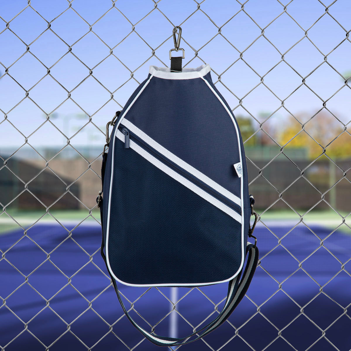 Navy blue sports pickleball  bag with white stripes hanging on a chain-link fence with a blurred tennis court background.
