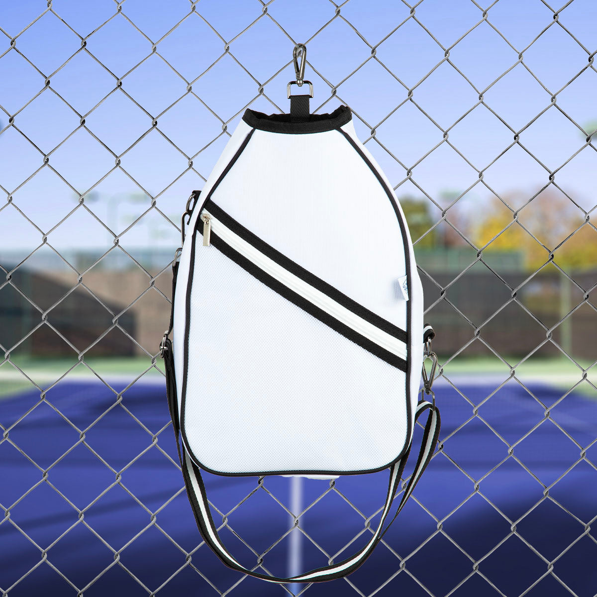 White sports  pickleball bag with black straps hanging on a chain-link fence with a blurred outdoor background.