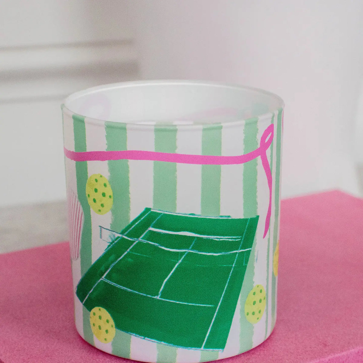 A frosted glass candle jar featuring a pickleball-themed illustration with green net and balls, yellow-green mops, and pink and green vertical stripes, resting on a pink surface.
