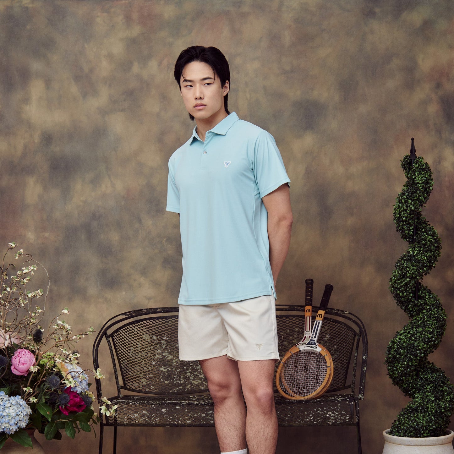 Man in light blue polo shirt and white shorts standing next to a chair with tennis rackets and decorative plants.