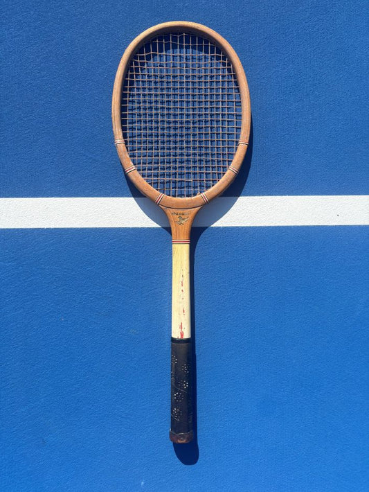 A vintage wooden tennis racket with a black grip, leaning against a blue tennis court wall with a white line.