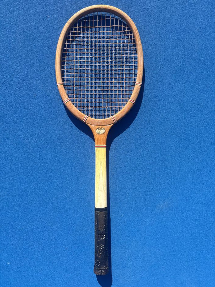 A vintage wooden tennis racket with a light-colored wooden shaft and a black grip, displayed vertically against a solid blue background.