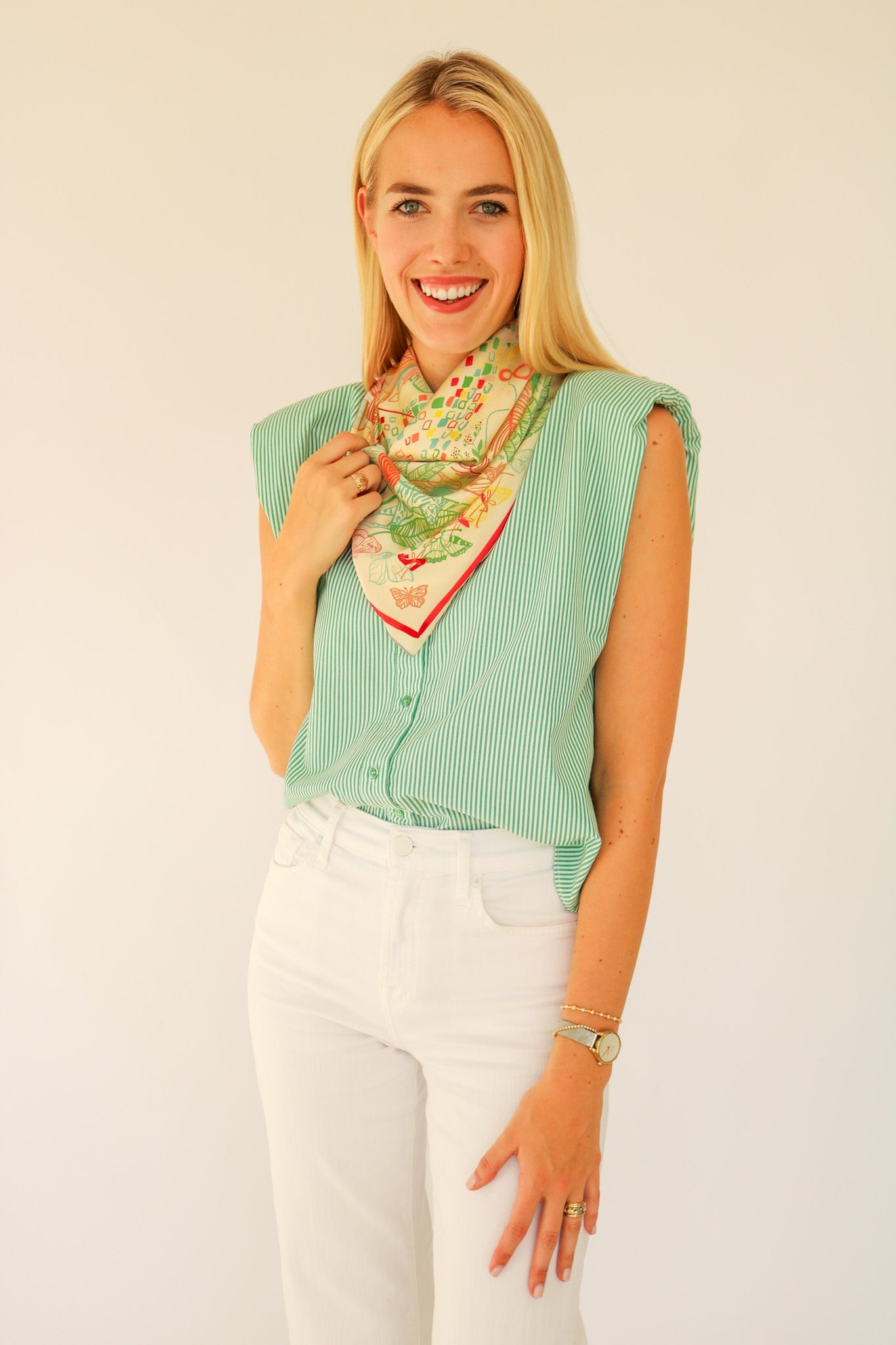 Woman wearing a green sleeveless top and white pants with a colorful scarf around her neck against a plain background