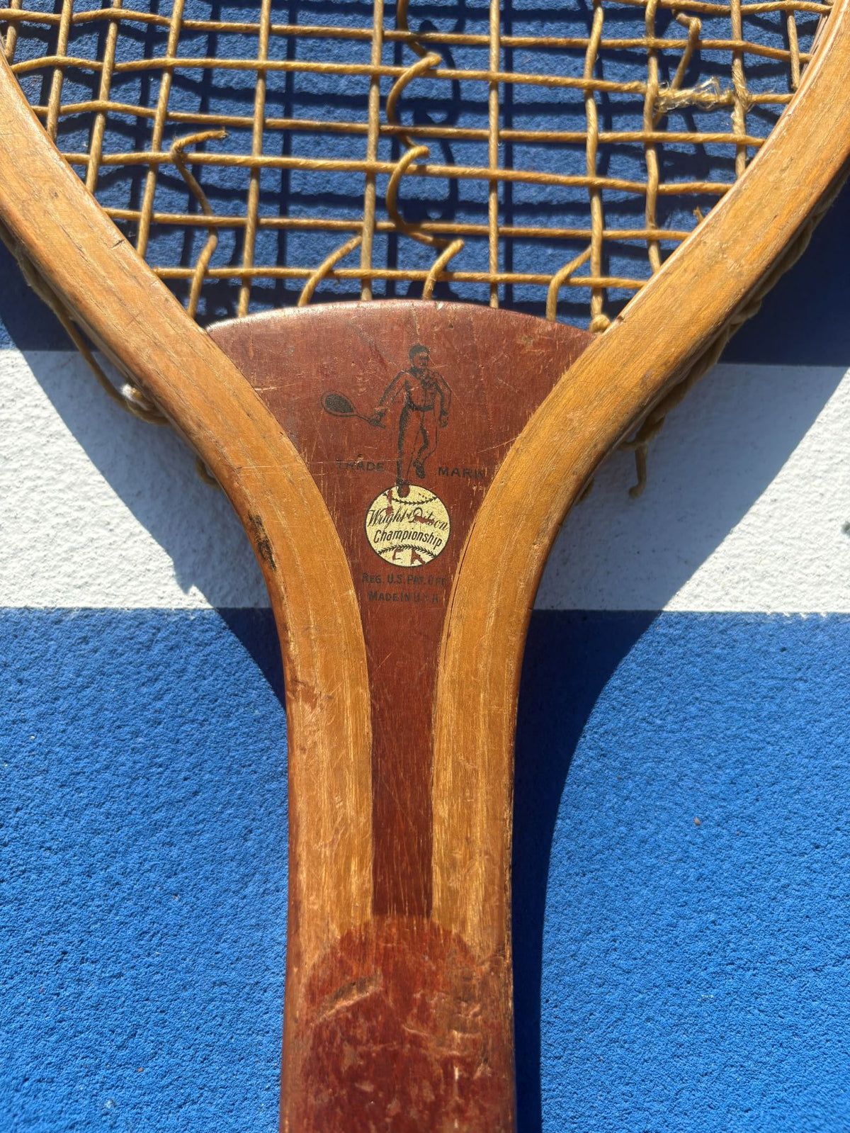 Close-up of a vintage wooden tennis racket head with a woven string bed, resting on a blue and white tennis court surface.