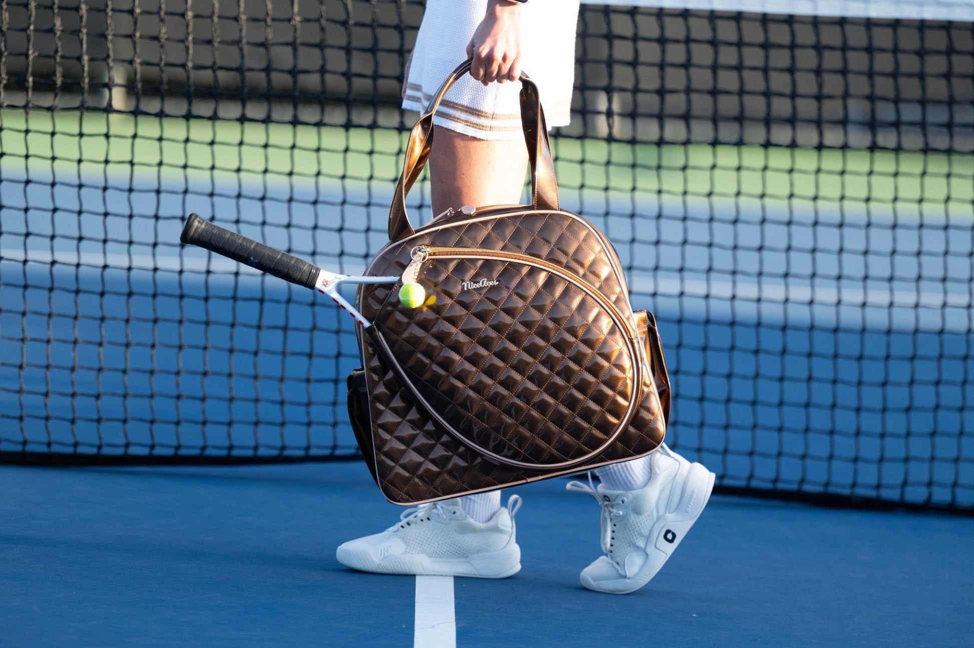 Person holding a brown quilted tennis bag with a racket on a tennis court