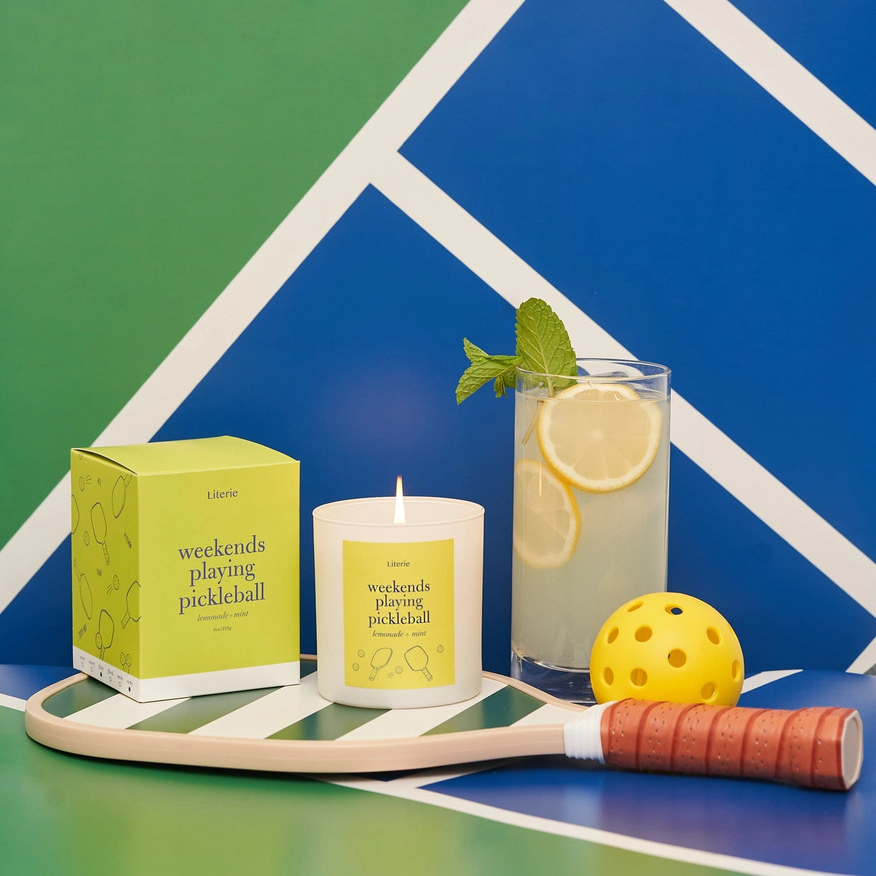 Candle with lemon and mint garnish, pickleball paddle, and ball on a colorful geometric background